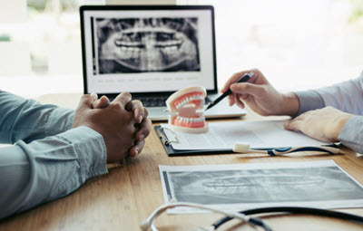 Dentist explaining dental X-rays and treatment plan using a teeth model, with a patient seated across the table in a consultation.