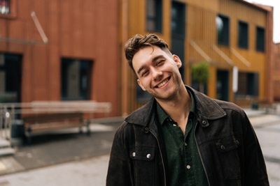 Smiling young man standing outdoors in front of modern buildings. He tilts his head slightly toward the camera.