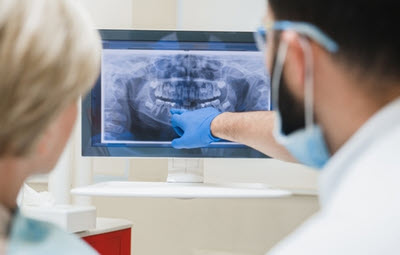 Dentist wearing gloves and a mask pointing at a dental X-ray on a monitor while discussing the image with a seated patient.